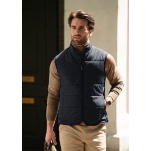 A man wearing a navy blue puffer vest over a tan sweater, walking outdoors. The vest has a zip closure and a logo.