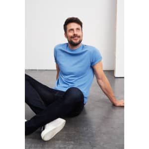 A blue T-shirt displayed on a seated male model wearing dark jeans and white sneakers, with a relaxed expression.