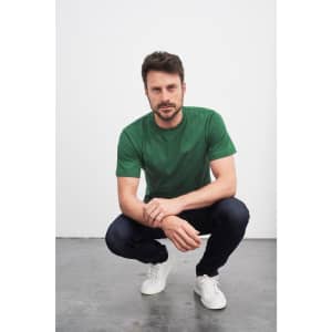 A green T-shirt worn by a man squatting in jeans and white sneakers, featuring a logo.