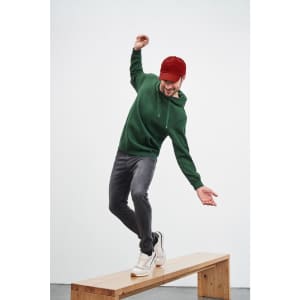 A man wearing a green hooded sweatshirt and a red cap balances on a wooden bench, dressed in grey jeans and white sneakers.