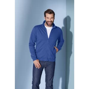 A blue track jacket with a zip, worn by a smiling man against a light background. Features a logo on the jacket.