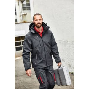 A man in a black softshell jacket carries a grey tool case, walking outdoors. The jacket features a hood and zippers.