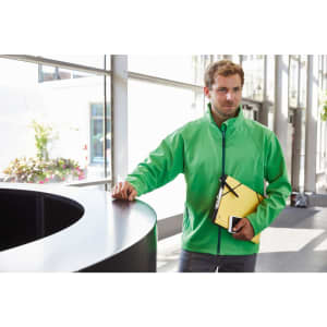 A man in a bright green softshell jacket stands near a black counter, holding a yellow folder and a notepad.