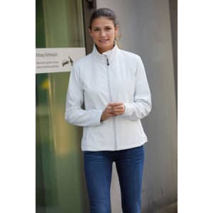 A woman wearing a white softshell jacket stands outdoors, smiling, with her hands in front of her.