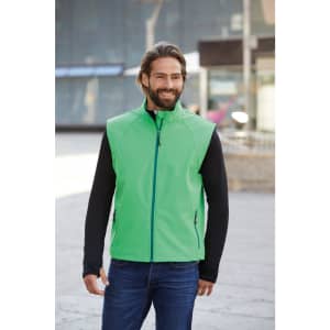 A green softshell vest with a zip front, paired with a black long-sleeve top, worn by a smiling man outdoors.