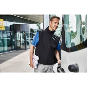 A man wearing a black softshell vest stands next to a vehicle, holding a clipboard. The vest has a logo.
