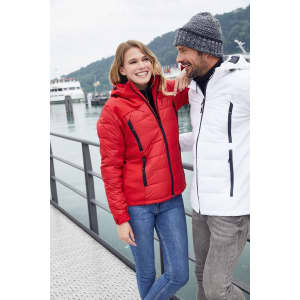 A red and a white soft shell jacket are displayed, worn by a couple against a scenic waterfront backdrop. The jackets have zippers and one features a logo.