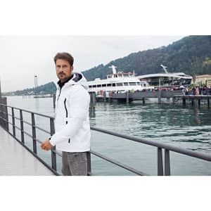 A man in a white soft shell jacket stands by a waterfront, with a boat and people in the background.
