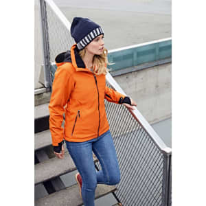 An orange soft shell jacket worn by a woman, paired with a blue beanie and denim jeans, standing on stairs.