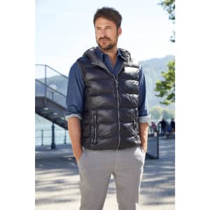 A man wearing a black down vest over a blue shirt and grey trousers, standing outdoors by a lakeside.