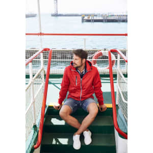A man in a red maritime jacket sits on green stairs by the sea, wearing shorts and white shoes. The jacket has a logo.
