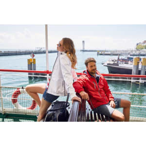 A man in a red maritime jacket and a woman in a white jacket pose on a boat near a harbour. The scene features water and a dock.