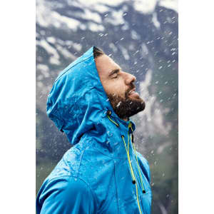 A blue men's outdoor jacket with a hood, worn by a man in the rain against a mountainous backdrop.