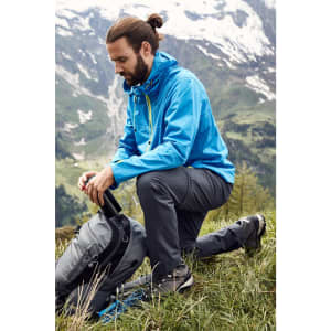 A blue men's outdoor jacket is worn by a person kneeling on grass next to a backpack in a mountainous landscape.