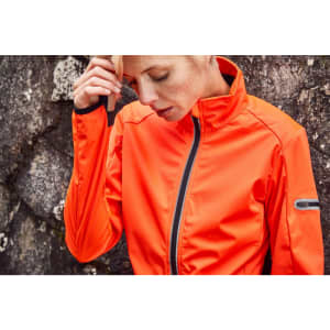 An orange ladies sports softshell jacket features a zip front and a logo, set against a textured rock background.