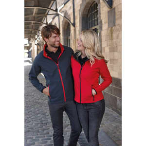 A grey and red softshell jacket is worn by a smiling couple in a cobblestone alley, with a building in the background.