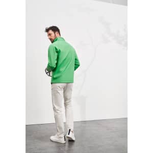 A man in a green softshell jacket stands against a white wall, wearing light trousers and holding a ball.