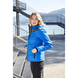 A blue puffer jacket with a hood displayed on a woman standing outdoors near a staircase and body of water.