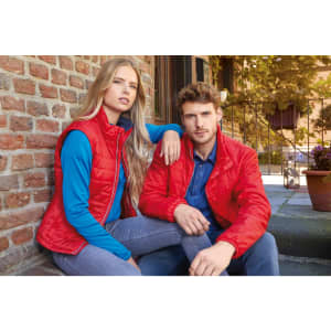 A red padded vest worn by a woman seated beside a man in a red jacket, both against a brick wall backdrop.