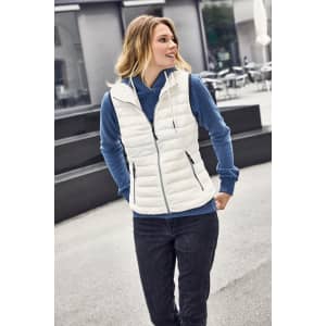A white down vest worn over a blue hoodie, displayed against an outdoor modern café setting. The vest features a front zip and pockets.