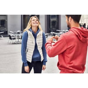 A smiling woman wears a white down vest over a blue hoodie, while a man in a red hoodie makes a heart shape with his hands.