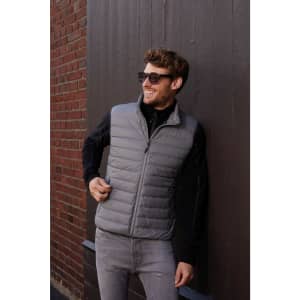 A man wearing a grey down vest stands against a brick wall, smiling and wearing sunglasses. The vest has a logo.