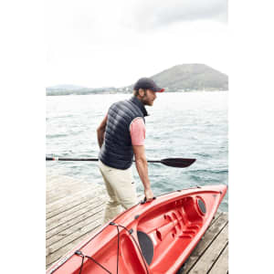 A man wearing a navy down vest and a cap, carrying a red kayak by a waterfront.