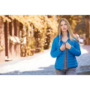 A blue ladies' down puffer jacket is worn by a model in an outdoor setting with autumn foliage.