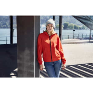A red ladies hooded softshell jacket worn by a woman leaning against a post, paired with a beanie and jeans.