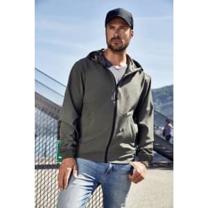 A man in a green hooded softshell jacket stands casually against an urban backdrop, wearing a cap and jeans.