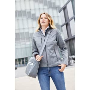 A grey ladies softshell jacket worn by a woman carrying a bag, set against a modern building backdrop.