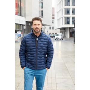 A man wearing a navy padded jacket stands on a city street. He has a beard and is dressed in blue trousers.
