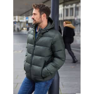 A green padded jacket with a hood worn by a man, featuring a puffer design and standing against a post in an urban setting.