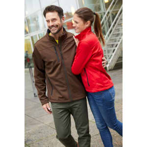 A red ladies softshell jacket is worn by a smiling woman alongside a man in a brown jacket. Both are outdoors.