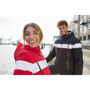 A red and black soft shell jacket with white stripes, worn by a smiling woman and a man by the water.