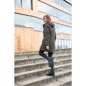 A woman wearing a green parka jacket with a hood, walking up outdoor steps. The jacket features a logo.