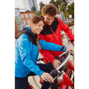 A blue and red ladies' all-weather jacket is worn by a woman and a man beside bicycles, set outdoors. The jackets have a logo.