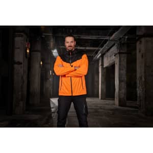 An orange and black men's allweather jacket is displayed on a model in an industrial setting. The jacket has a logo.