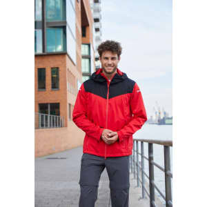 A man wearing a red and black all-weather jacket, standing near a waterfront with buildings in the background.
