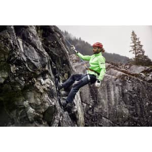 A man in a bright green jacket and climbing gear descends a rocky cliff, wearing black zip-off trekking pants.