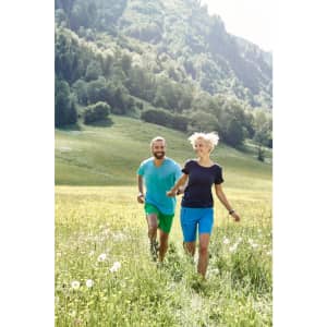 A couple running in a grassy field, both wearing bright blue and green trekking shorts. The landscape features lush green hills.