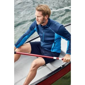 A man in a blue zip-up top and navy shorts sits in a rowing boat, holding a red paddle against a water backdrop.