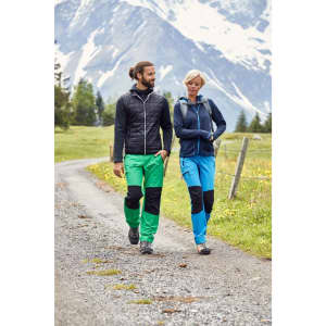 Two people walking on a trail, wearing green and blue trekking pants with black patches, set against a mountainous background.
