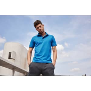 A man wearing a blue mercerised polo shirt stands outdoors, hands in pockets, against a cloudy sky backdrop.