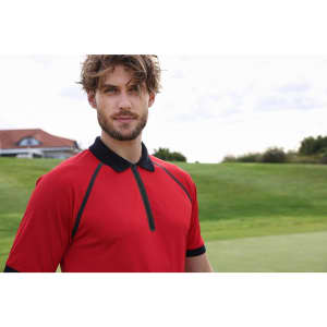 A man in a red zip polo shirt with black accents stands on a golf course background. The shirt features a collar and a logo.