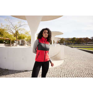 A fleece vest in red and grey, featuring zippered pockets and a logo, worn by a model outdoors.