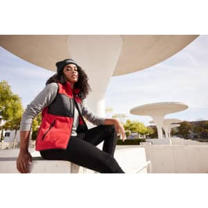 A woman is wearing a red and black fleece vest with a grey long-sleeve top, seated outdoors against a modern backdrop.