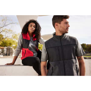 A grey and black men's fleece vest is worn by a man, while a woman in a red and black vest sits nearby.