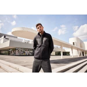 A mens fleece jacket in black, worn by a model standing outdoors near modern architecture. The jacket features a logo.