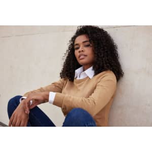 A knitted round-neck pullover in tan, featuring a woman sitting against a wall, wearing a collared shirt underneath.